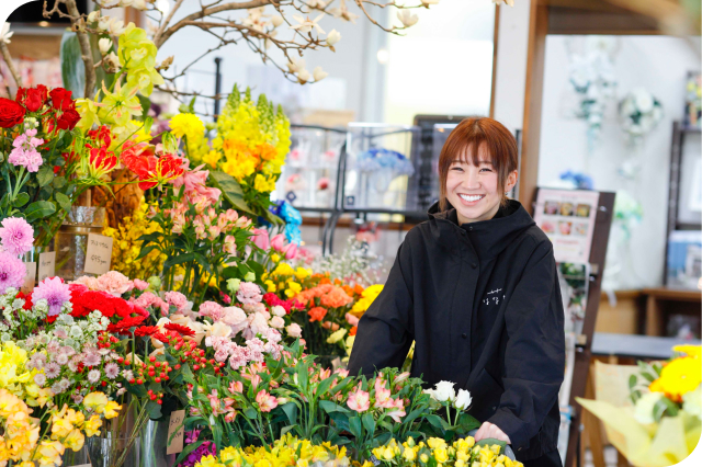 色とりどりの花が並ぶ生花店で、黒い上着を着た女性店員がカメラに向かって満面の笑みを浮かべている。背景にはバラやダリア、黄色い花など多様な花が豊富に陳列されている。