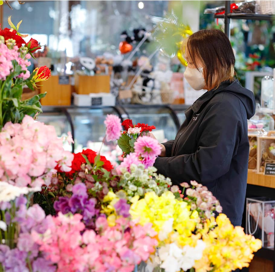 花屋の店内で青い花を選ぶマスク姿の女性と、それを見守る別の女性。