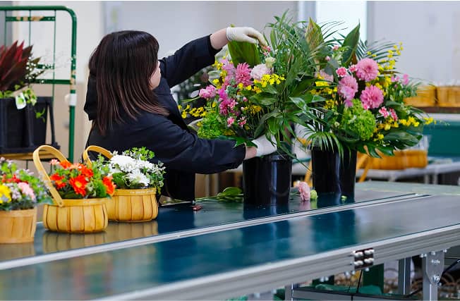 コンベアライン上で大型のフラワーアレンジメントを整える作業員、色鮮やかな花々が並ぶ