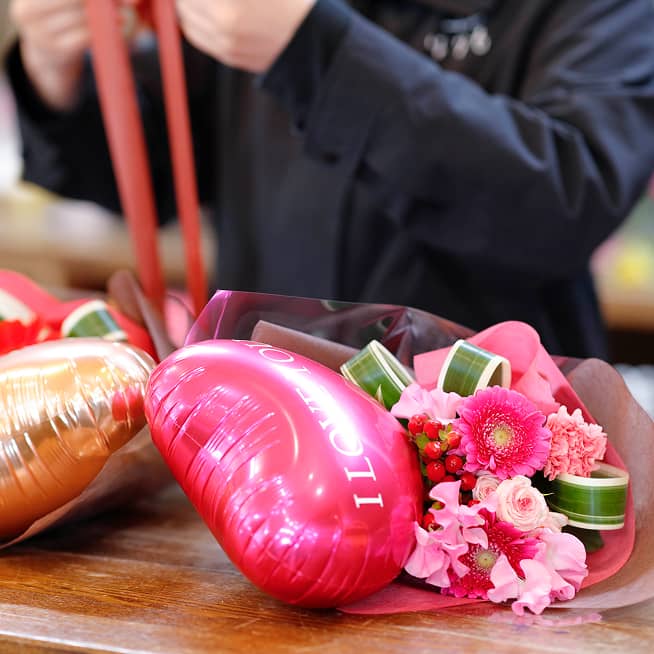 『はなも』のスタッフがピンクのハート型バルーンと花を組み合わせた花束を丁寧にラッピングしている様子