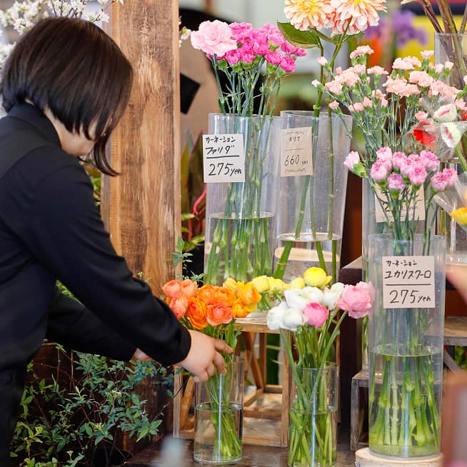 黒い服を着た人物が、様々な色の花が飾られた店内で、ピンク色のカーネーションが活けられた花瓶を調整している。値札には「カーネーション」「¥275」と書かれている。