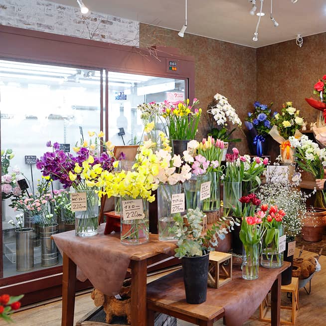 花屋の店内で、たくさんの花瓶に様々な色の花が活けられている。店内のテーブルには、黄色のオンシジュームやピンクのチューリップなど、色鮮やかな花が並べられている。