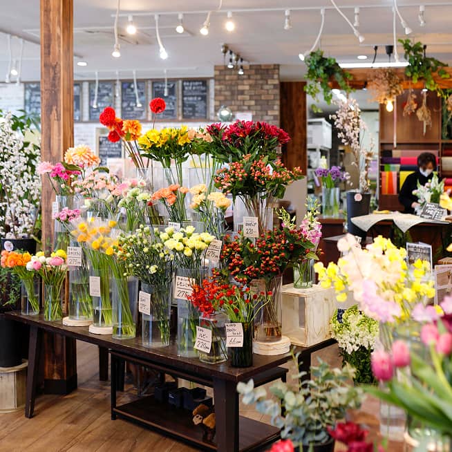 花屋の店内には、カラフルな花が活けられた多数の花瓶が木製のテーブルに並べられている。店内の奥には、黒い服を着てマスクを着用した人物がいる。