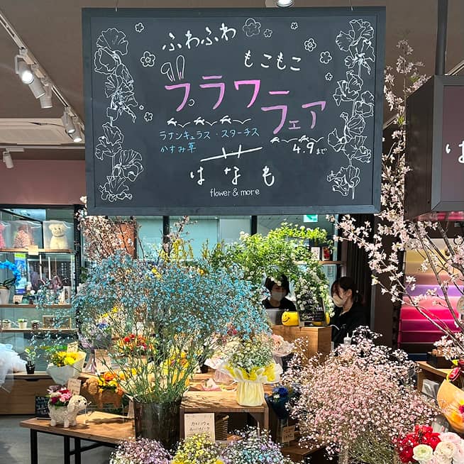 ふわふわ」「もこもこ」「フラワーフェア」「はなも flower & more」と書かれた黒い看板の下に、色とりどりの花が飾られた花屋の様子。特に、青いカスミソウが目を引く。
