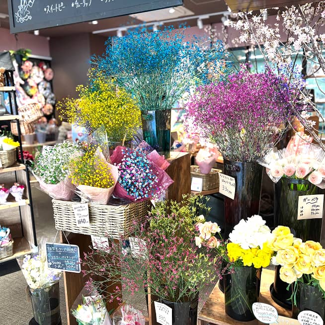 青、黄、紫など様々な色に染められたカスミソウが目を引く花屋の店内。木製のカゴやガラスの花瓶に活けられた花が並ぶ。